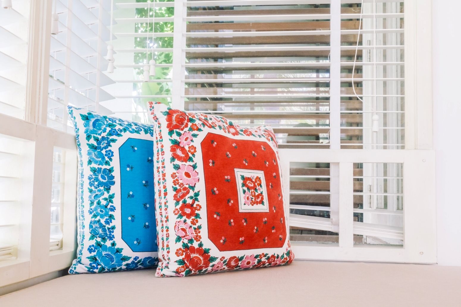 Window Treatment Design 1 Decorative floral pillows on a windowsill with white blinds; blue and red patterns showcase bold floral prints.