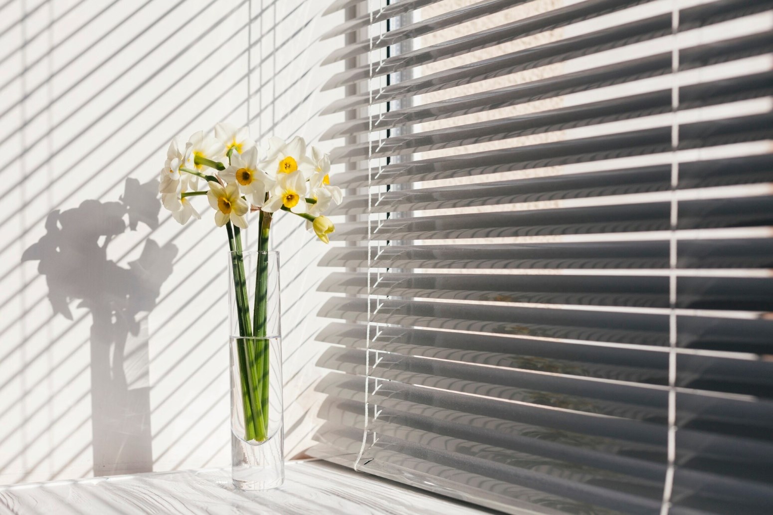 Window Treatment Design 2 - Royal Crest, Inc. White daffodils in a tall glass vase on a windowsill, with striped blinds casting shadows on the wall.